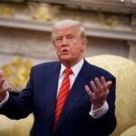 A man in a suit speaking with expressive hand gestures in a formal setting