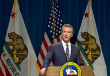 Newsom’s Ex-Chief ARRESTED – Massive Fraud Uncovered Man speaking at podium with California and US flags