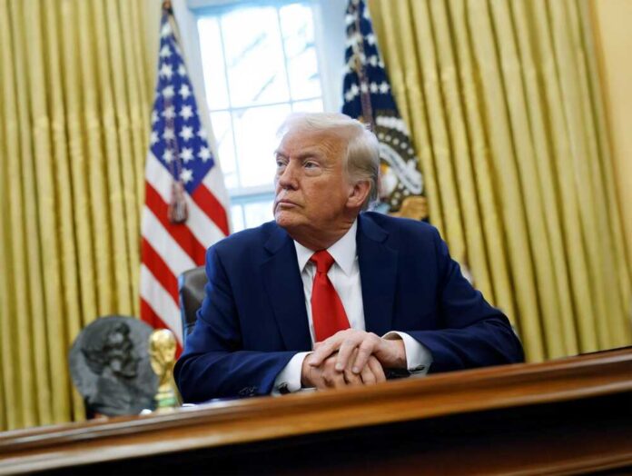 A man in a suit sitting at a desk in the Oval Office with American flags in the background